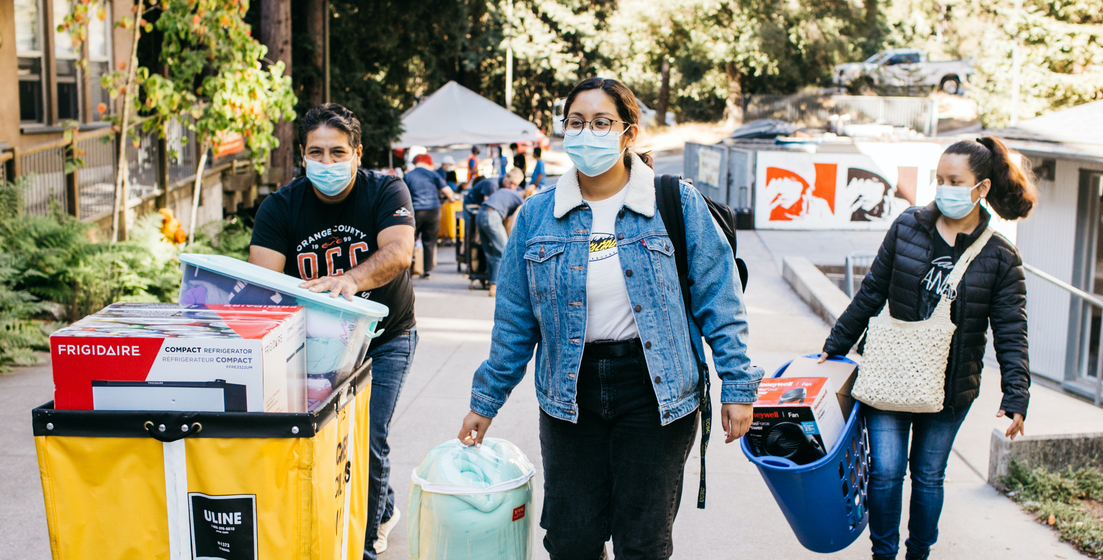 A family of three walks towards the camera. All three are wearing blue medical masks and are carrying a variety of belongings. The person on the left is pushing a yellow cart.
